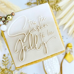 Op de afbeelding: Een vierkante koekje versierd met witte glazuur en de woorden "'Tis the season to be jolly" in cursief schrift. Het koekje is bedekt met gouden en witte sprinkles.