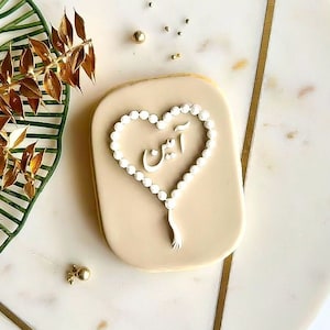 May include: A rectangular cookie with a light beige glaze. A white beaded heart design with the word "آمين" in the center. Gold leaf and decorative elements are in the background.