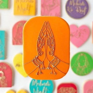 May include: Orange cookie with a gold design of two hands in a prayer position. The hands have intricate henna designs and gold bracelets.