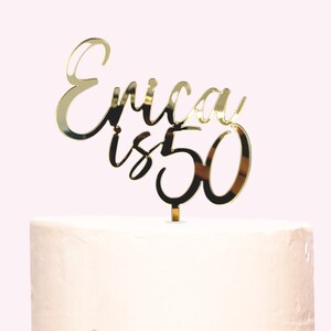 May include: Gold mirror acrylic cake topper with the text "Erica is 50" in a cursive font.