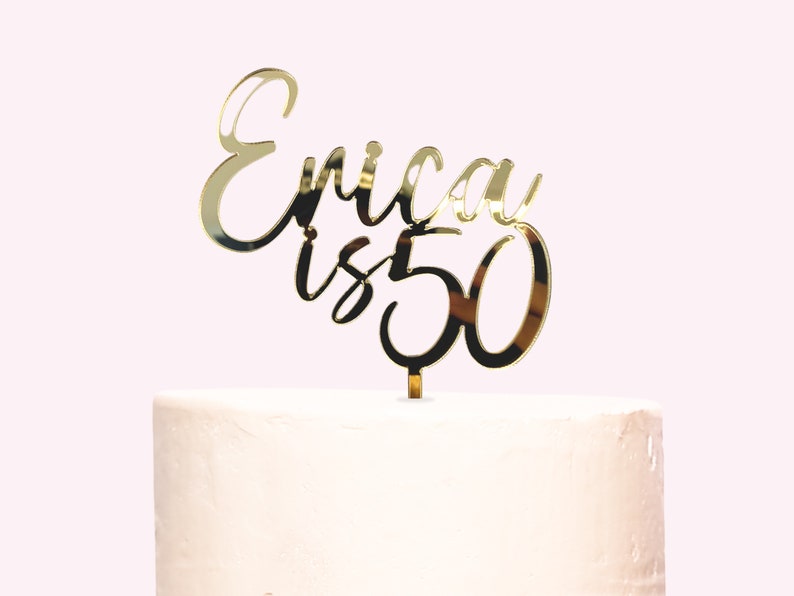 May include: Gold mirror acrylic cake topper with the text "Erica is 50" in a cursive font.