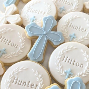 Puede incluir: Una colección de galletas de azúcar decoradas con glaseado azul y blanco. Las galletas presentan los nombres "Hilton" y "Hunter" y un diseño de cruz.