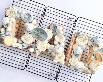 Könnte beinhalten: Ein Kuchen in Form des Wortes "DAD", verziert mit weißem, hellblauem und cremefarbenem Zuckerguss. Der Kuchen steht auf einem schwarzen Drahtgestell und enthält ein kleines Oval mit dem Text "Happy Father's Day".
