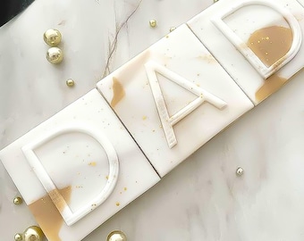 May include: Four white square tiles with the word "DAD" spelled out in raised letters. The tiles have a light brown marble pattern and gold glitter accents.