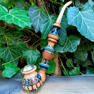 May include: A handcrafted wooden tobacco pipe with a curved stem. The pipe features intricate geometric patterns in blue, brown, and yellow. The bowl has a lid, and the stem is light-coloured. The pipe is set against a backdrop of green ivy.