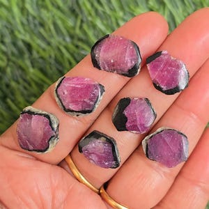 Op de afbeelding: Een verzameling ruwe, natuurlijke edelstenen. De stenen hebben een levendige roze kleur met een zwarte rand. De edelstenen worden op een hand gepresenteerd, tegen een groene achtergrond.