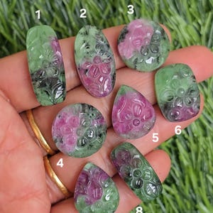 May include: A selection of carved gemstones in diverse shapes, such as oval, teardrop, and round. The stones display green and pink colours, with floral carvings. The gemstones are held in a hand against a backdrop of green grass.