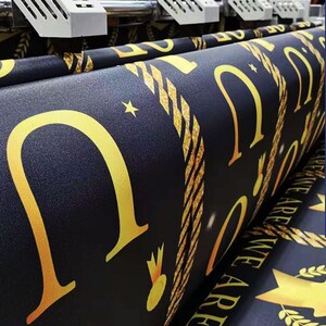 Puede incluir: Primer plano de una impresora de gran formato imprimiendo una pancarta negra y dorada con el texto "Class of 2021 We Are The Future". La pancarta se est&aacute; imprimiendo en un rollo de tela.