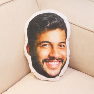 May include: A white cushion with a printed image of a person's face. The person is smiling and has short brown hair. The cushion is on a white sofa.
