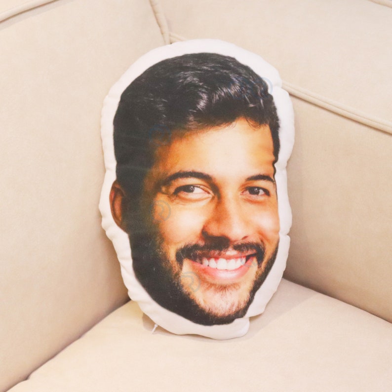 May include: A white cushion with a printed image of a person's face. The person is smiling and has short brown hair. The cushion is on a white sofa.