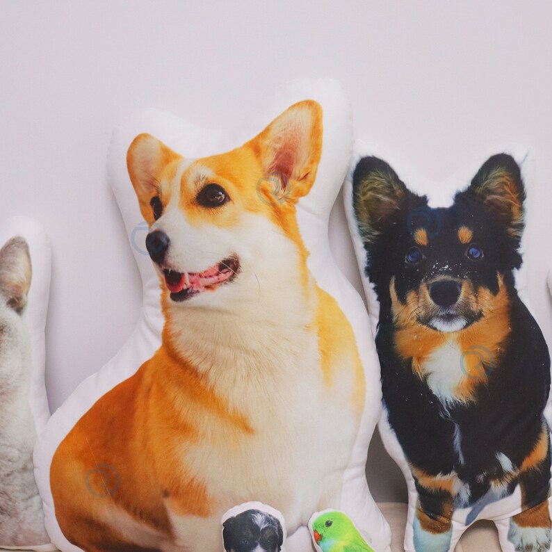 May include: Three plush dog cushions, one featuring a corgi, one with a black and brown dog, and one with a black and white dog. The cushions are all white with the dog images printed on them.