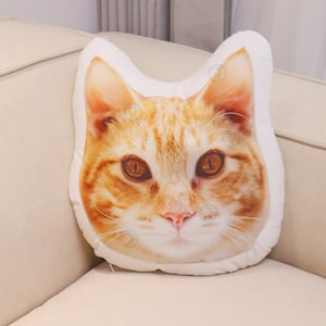 Op de afbeelding: Een wit kussen met een bedrukte afbeelding van een kattenkop. De kat heeft oranje en wit haar en bruine ogen.