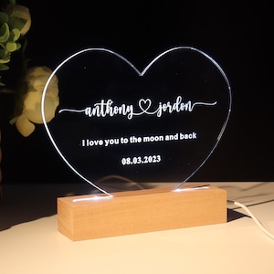 Könnte beinhalten: Ein herzförmiges Acryl-Nachtlicht mit einem Holzsockel. Das Licht zeigt die Namen "anthony & jordan" und den Text "Ich liebe dich bis zum Mond und zurück" mit dem Datum "08.03.2023".