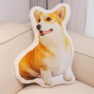 Peut inclure: Un oreiller en peluche blanc en forme de corgi. L'oreiller est imprim&eacute; d'une image r&eacute;aliste d'un corgi, avec une fourrure brune et blanche, un nez noir et une langue rose.
