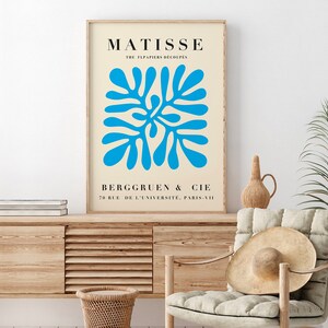 Op de afbeelding: Een ingelijste kunstdruk met een blauw abstract ontwerp op een cr&egrave;mekleurige achtergrond. De print bevat de naam "MATISSE" en de tekst "THE FLPAPIERS DECOUPES" bovenaan, met "BERGGRUEN & CIE" en adresgegevens eronder. Het kunstwerk is ingelijst in een licht houten frame.