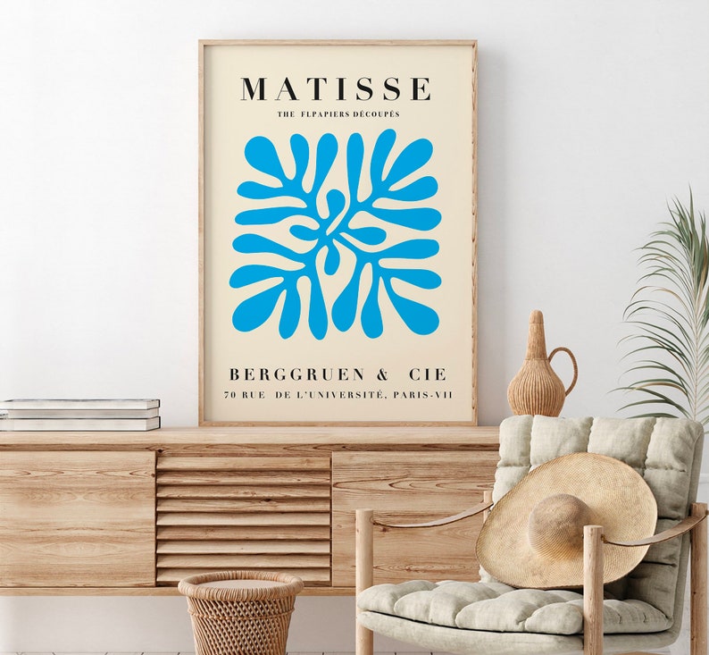 Op de afbeelding: Een ingelijste kunstdruk met een blauw abstract ontwerp op een cr&egrave;mekleurige achtergrond. De print bevat de naam "MATISSE" en de tekst "THE FLPAPIERS DECOUPES" bovenaan, met "BERGGRUEN & CIE" en adresgegevens eronder. Het kunstwerk is ingelijst in een licht houten frame.
