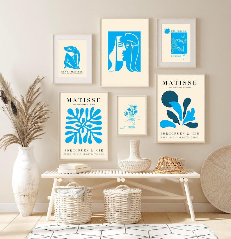 Op de afbeelding: Een galerijmuur met ingelijste kunstprints in blauwe en cr&egrave;mekleuren. De prints tonen abstracte ontwerpen, bloemmotieven en tekst, waaronder "HENRI MATISSE" en "BERGGRUEN & CIE". De kunstwerken worden tentoongesteld boven een witte bank met geweven manden.