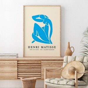 Op de afbeelding: Een ingelijste kunstdruk met een blauwe Matisse-uitgesneden figuur op een cr&egrave;mekleurige achtergrond. De print bevat de tekst "HENRI MATISSE" en "THE CUT OUTS - 1952 GALERIE MAEGHT". Het kunstwerk wordt gepresenteerd in een licht houten frame.