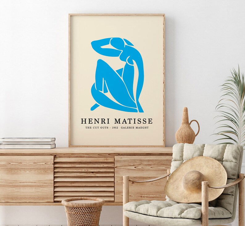 Op de afbeelding: Een ingelijste kunstdruk met een blauwe Matisse-uitgesneden figuur op een cr&egrave;mekleurige achtergrond. De print bevat de tekst "HENRI MATISSE" en "THE CUT OUTS - 1952 GALERIE MAEGHT". Het kunstwerk wordt gepresenteerd in een licht houten frame.