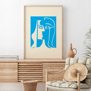 Op de afbeelding: Een ingelijste kunstdruk met een blauw en wit abstract portret. Het kunstwerk, gesigneerd door Picasso, toont het profiel van een vrouw met gedurfde, geometrische vormen. De print wordt gepresenteerd op een lichtbeige achtergrond en voegt een vleugje moderne kunst toe.