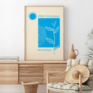 Op de afbeelding: Een ingelijste kunstdruk met een blauw en wit botanisch ontwerp op een cr&egrave;mekleurige achtergrond. Het kunstwerk bevat een blauwe vierkant met een zonachtig patroon, een cirkel en het woord "BOH&Eacute;ME" in blauw. De print wordt getoond op een houten kast.
