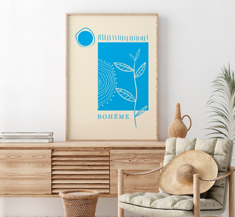 Op de afbeelding: Een ingelijste kunstdruk met een blauw en wit botanisch ontwerp op een cr&egrave;mekleurige achtergrond. Het kunstwerk bevat een blauwe vierkant met een zonachtig patroon, een cirkel en het woord "BOH&Eacute;ME" in blauw. De print wordt getoond op een houten kast.