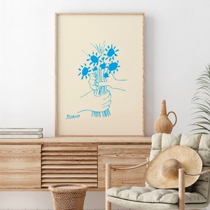Op de afbeelding: Een ingelijste kunstdruk met een blauwe lijntekening van een boeket bloemen, vastgehouden door handen. Het kunstwerk staat op een cr&egrave;mekleurige achtergrond en is gesigneerd met "Picasso". De lijst is van licht hout.