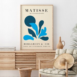 Op de afbeelding: Een ingelijste kunstprint met een Matisse-ontwerp, een beige achtergrond en blauwe abstracte vormen. De print bevat de naam "MATISSE" en de tekst "THE FLPAPIERS DECOUPES" bovenaan, en "BERGGRUEN & CIE" en een adres onderaan.