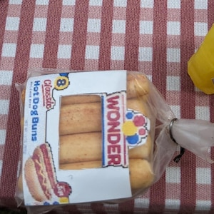 May include: A pack of eight classic hot dog buns, branded as Wonder, alongside a yellow mustard bottle. The buns are in a clear plastic bag, placed on a red and white checkered tablecloth.