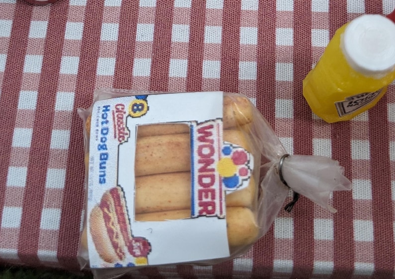May include: A pack of eight classic hot dog buns, branded as Wonder, alongside a yellow mustard bottle. The buns are in a clear plastic bag, placed on a red and white checkered tablecloth.