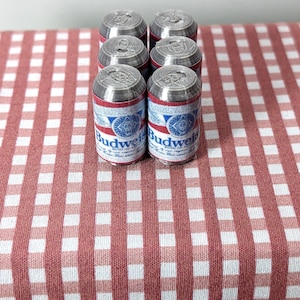 May include: Six miniature Budweiser beer cans with red, white, and blue labels are arranged on a red and white checked tablecloth. The cans are silver on top and feature the Budweiser logo.