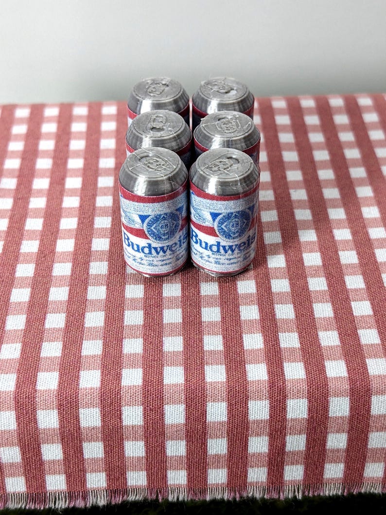 May include: Six miniature Budweiser beer cans with red, white, and blue labels are arranged on a red and white checked tablecloth. The cans are silver on top and feature the Budweiser logo.