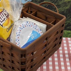 May include: A brown woven picnic basket containing snacks and plates. Inside are a bag of crisps, paper plates with red and blue stars, and napkins. The basket rests on a red and white checked tablecloth.