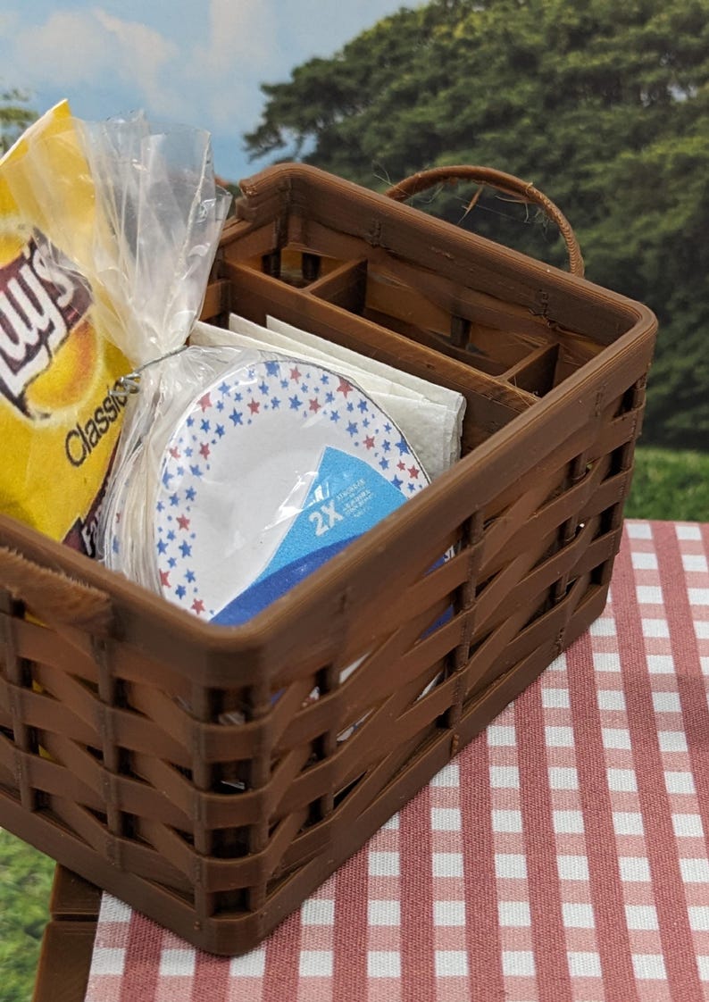 May include: A brown woven picnic basket containing snacks and plates. Inside are a bag of crisps, paper plates with red and blue stars, and napkins. The basket rests on a red and white checked tablecloth.
