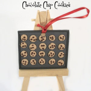 May include: A miniature baking sheet with 20 chocolate chip cookies on it. The baking sheet is on a small wooden easel with a red ribbon attached to the top.