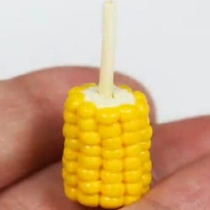 May include: A miniature, bright yellow corn on the cob with a light-coloured wooden stick inserted into the top. The corn kernels are detailed and arranged in neat rows. The miniature food item is held in a hand.