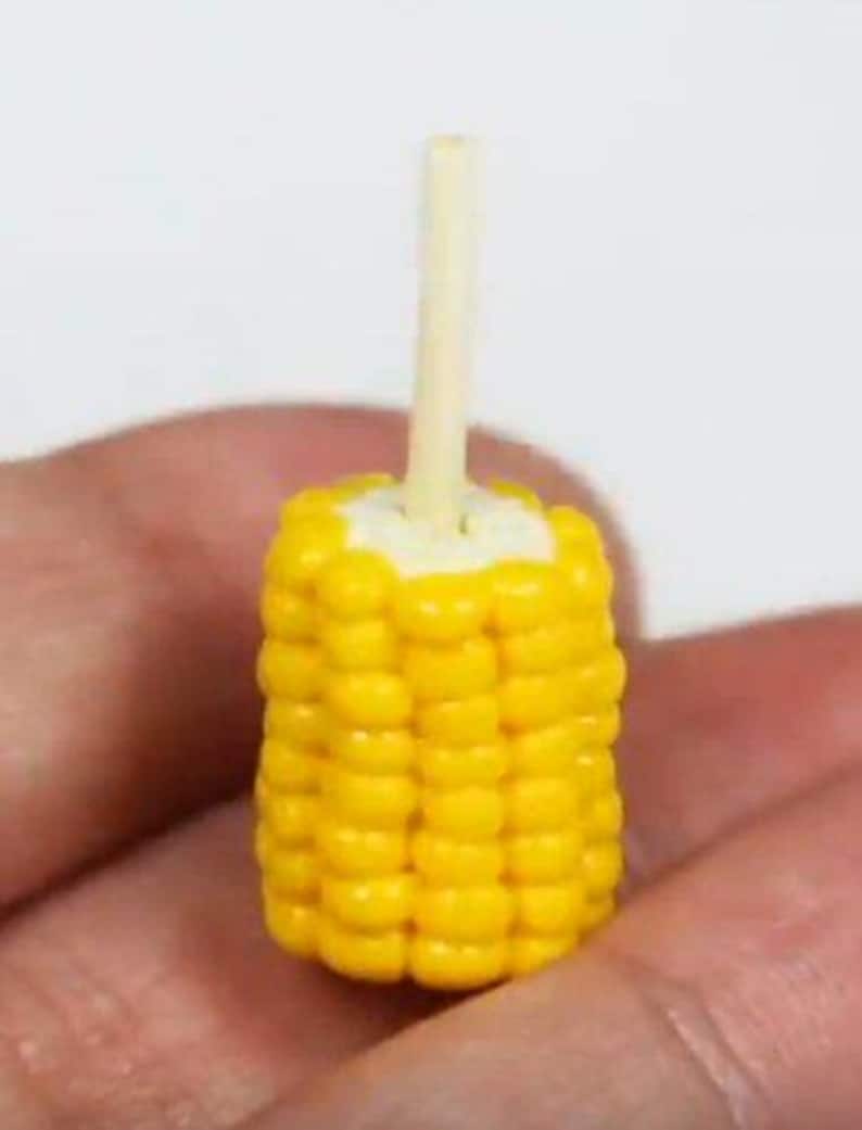 May include: A miniature, bright yellow corn on the cob with a light-coloured wooden stick inserted into the top. The corn kernels are detailed and arranged in neat rows. The miniature food item is held in a hand.