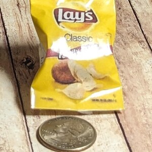May include: A yellow packet of Lay's Classic potato crisps with a quarter coin beside it. The packet displays the Lay's logo and the word "Classic". The crisps are visible through the packet.