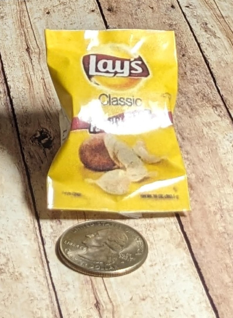 May include: A yellow packet of Lay's Classic potato crisps with a quarter coin beside it. The packet displays the Lay's logo and the word "Classic". The crisps are visible through the packet.