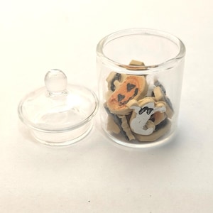 以下が含まれることがあります： ハロウィーンをテーマにしたミニチュアのクッキーが入った、蓋付きの透明なガラス瓶。クッキーには、ジャック・オー・ランタンと幽霊のデザインが施されています。瓶は開いており、蓋は白い表面の横に置かれています。