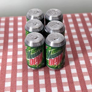 May include: Six miniature Mountain Dew cans, green and red, are arranged on a red and white checked tablecloth. The cans have silver tops and the Mountain Dew logo is visible.