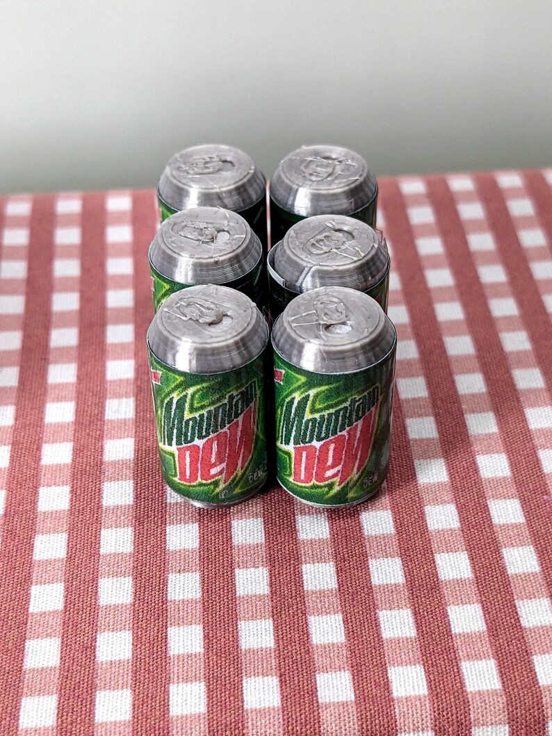 May include: Six miniature Mountain Dew cans, green and red, are arranged on a red and white checked tablecloth. The cans have silver tops and the Mountain Dew logo is visible.