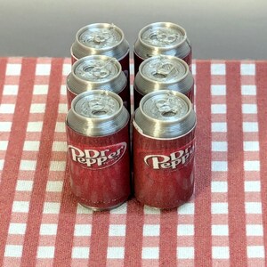 May include: Six miniature Dr Pepper soda cans with silver tops and red and white labels. The cans are arranged in a group on a red and white checkered surface. The Dr Pepper logo is visible on the cans.