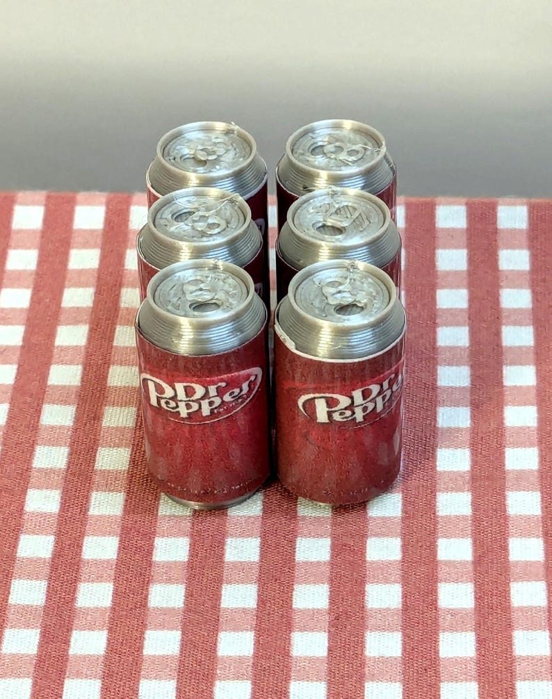 May include: Six miniature Dr Pepper soda cans with silver tops and red and white labels. The cans are arranged in a group on a red and white checkered surface. The Dr Pepper logo is visible on the cans.