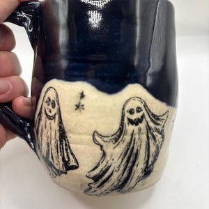 May include: A ceramic mug featuring a two-tone design with a glossy dark blue upper section and a speckled cream-colored lower section. The cream area is decorated with two black ink-style illustrations of ghosts and a small star symbol.