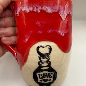 May include: A ceramic mug with a glossy red glaze dripping over a speckled cream-colored base. The front features a black ink illustration of a potion bottle topped with a heart, with the word LOVE printed on the bottle body.