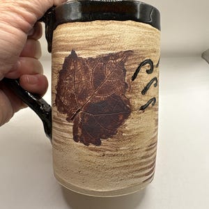 May include: A cylindrical ceramic mug with a textured beige body featuring a dark brown embossed leaf imprint and a glossy black rim and handle. The surface has horizontal brushstroke patterns in light brown, and the mug is held by a handle.