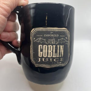 May include: Black ceramic mug with a white base, featuring a label that reads "IMPORTED 100% PURE GOBLIN JUICE" in a vintage style. The mug has a curved handle and is held by a hand.