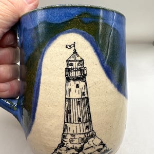 May include: A ceramic mug featuring a hand-drawn black ink illustration of a lighthouse on a cream-colored background. The mug has a glossy blue and green glaze that frames the central lighthouse design, with a handle on the left side.