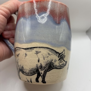 May include: A ceramic mug featuring a hand-drawn black ink illustration of a pig on a cream-colored base. The upper portion of the mug has a smooth, glossy glaze transition from a soft lavender-blue to a warm, reddish-orange at the rim.